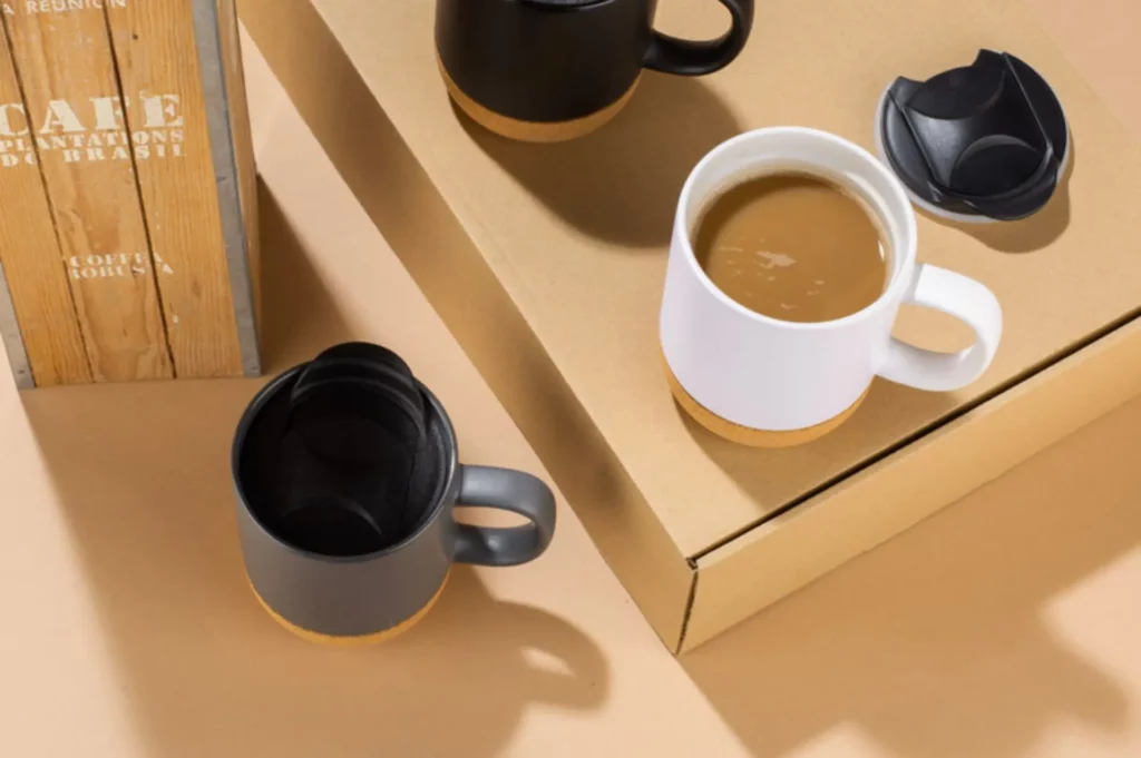 Trei căni ceramice personalizate Softel - albă, gri și neagră - cu bază de plută, aranjate pe cutii de carton, reprezentând un cadou corporate ideal.